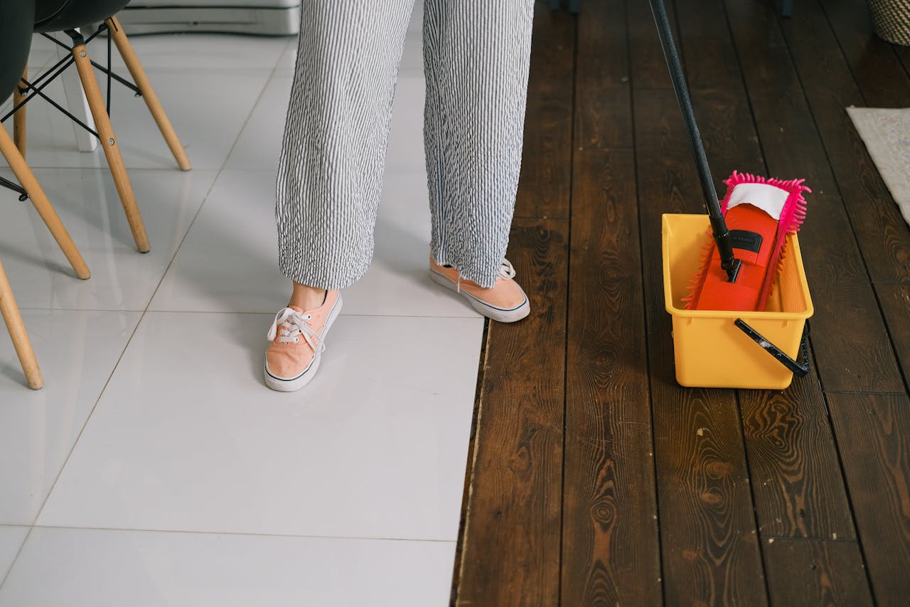 Crop anonymous woman in gumshoes and pants dipping mop into bucket with water on parquet in house